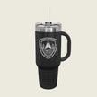 A matte black insulated travel tumbler with a clear lid and straw, featuring a white shield logo on the front with a skull and bridge graphic, set against a solid light beige background.
