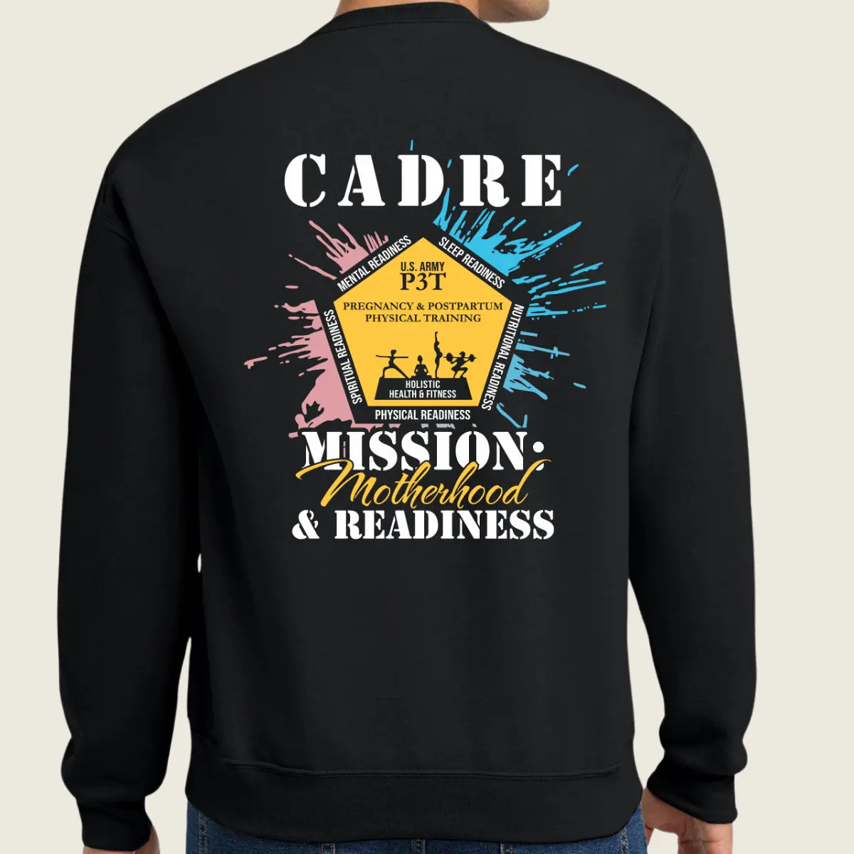 A person shown from the back wearing a black crewneck sweatshirt with a large graphic print. The graphic features the text 'CADRE' and 'MISSION Motherhood & READINESS' in white and yellow, surrounded by a yellow badge design with pink and blue paint splatters on a light beige background.