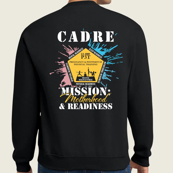 A person shown from the back wearing a black crewneck sweatshirt with a large graphic print. The graphic features the text 'CADRE' and 'MISSION Motherhood & READINESS' in white and yellow, surrounded by a yellow badge design with pink and blue paint splatters on a light beige background.