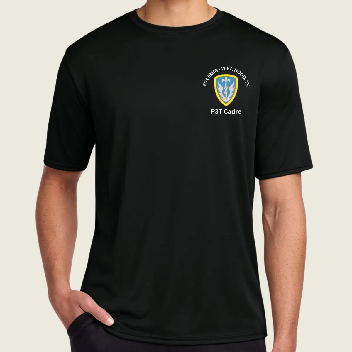 A man wearing a black short-sleeve crewneck t-shirt with a small military-style crest on the left chest. The crest features a yellow and blue shield with white text reading Cadre'.