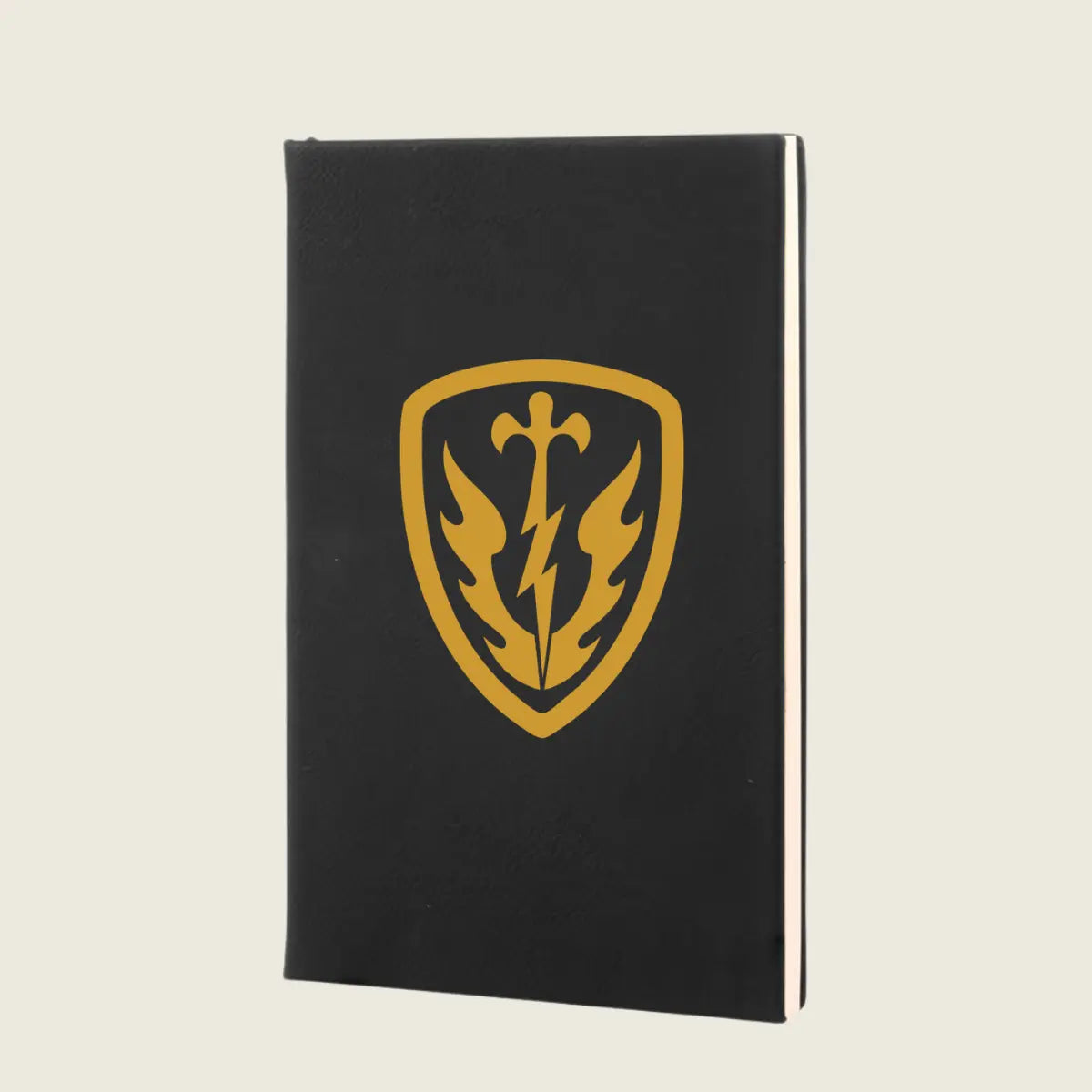 A black hardcover notebook featuring a gold shield emblem with a sword, lightning bolt, and flame design on the front cover, set against a solid light beige background.