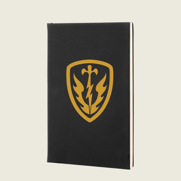 A black hardcover notebook featuring a gold shield emblem with a sword, lightning bolt, and flame design on the front cover, set against a solid light beige background.