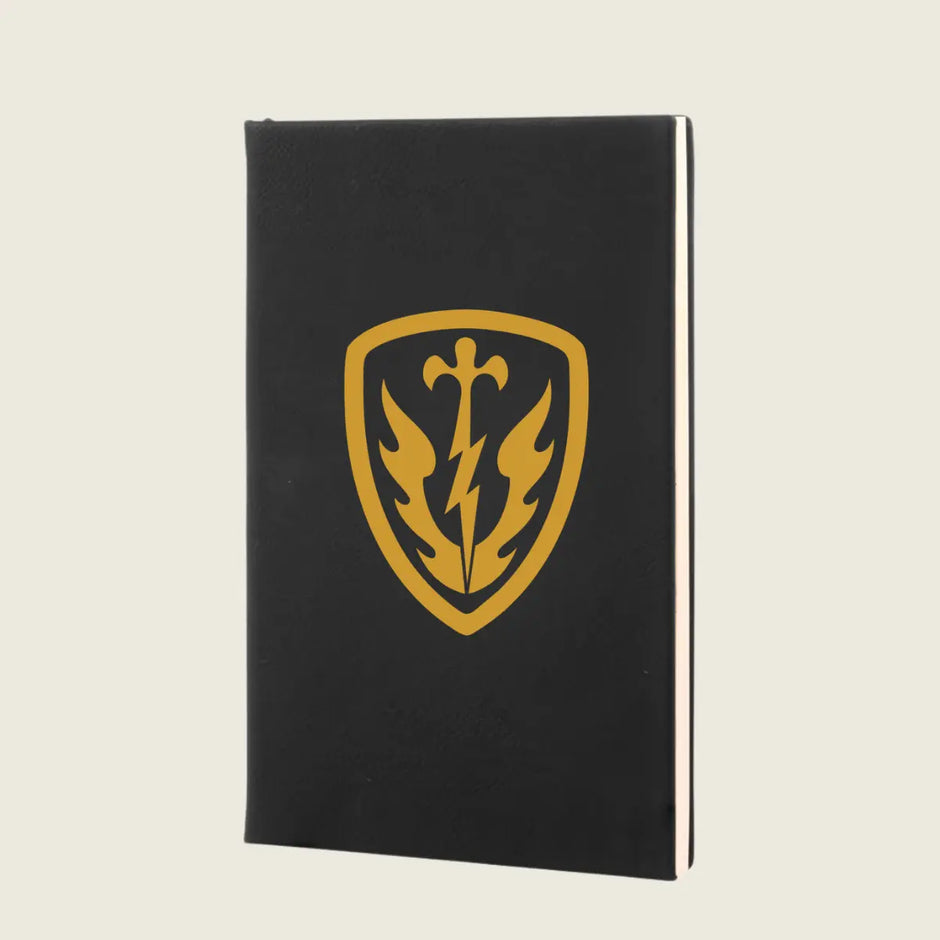 A black hardcover notebook featuring a gold shield emblem with a sword, lightning bolt, and flame design on the front cover, set against a solid light beige background.