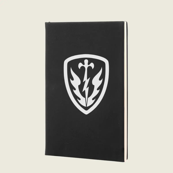 A black hardcover notebook or journal standing upright, featuring a white shield emblem on the front cover with a sword, lightning bolt, and winged design, set against a light beige background.