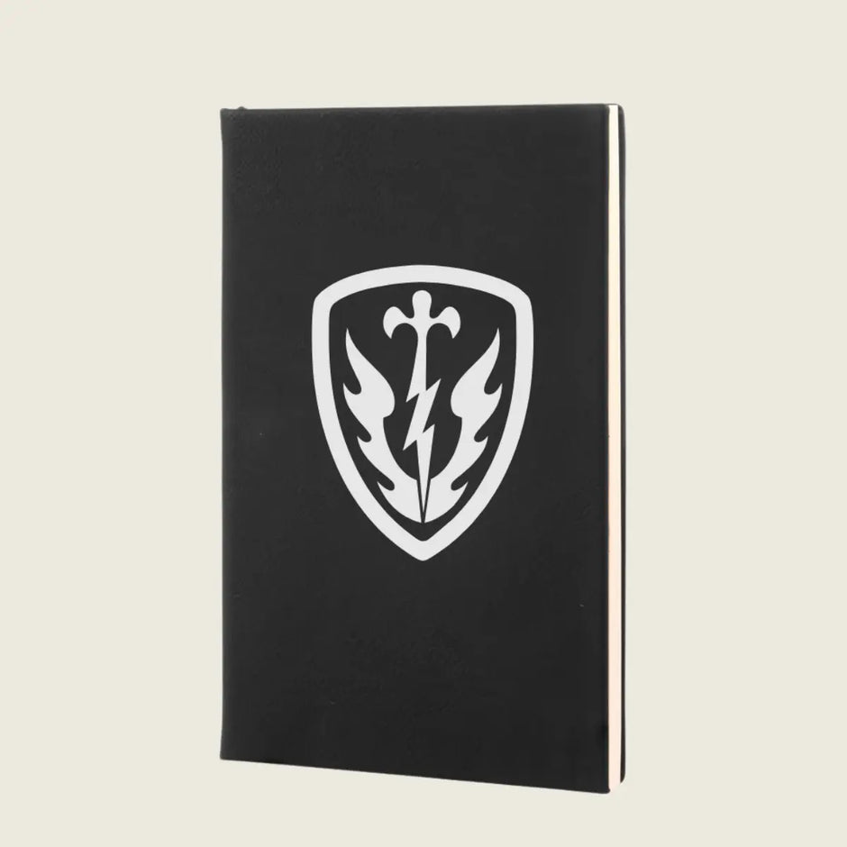 A black hardcover notebook or journal standing upright, featuring a white shield emblem on the front cover with a sword, lightning bolt, and winged design, set against a light beige background.