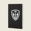 A black hardcover notebook or journal standing upright, featuring a white shield emblem on the front cover with a sword, lightning bolt, and winged design, set against a light beige background.