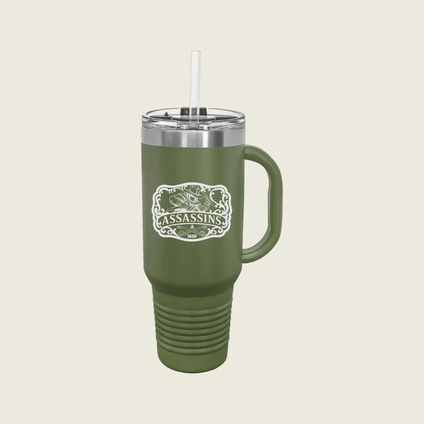 An olive green insulated tumbler with a handle, clear lid, and white straw, featuring a white and green 'Assassins' logo on the front, set against a solid light beige background.