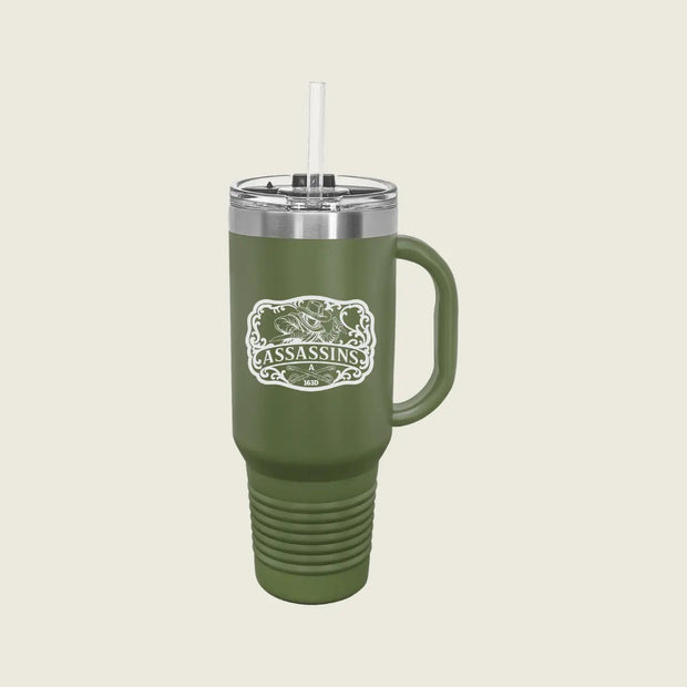 An olive green insulated tumbler with a handle, clear lid, and white straw, featuring a white and green 'Assassins' logo on the front, set against a solid light beige background.