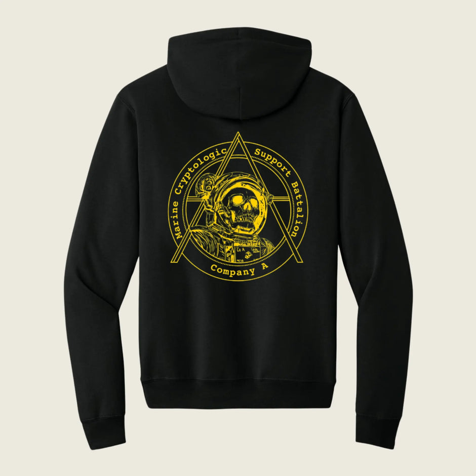 A black pullover hoodie shown from the back, featuring a large yellow graphic print of an astronaut inside a triangle emblem with the text 'Marine Cryptologic Support Mission Company A' on a light beige background.