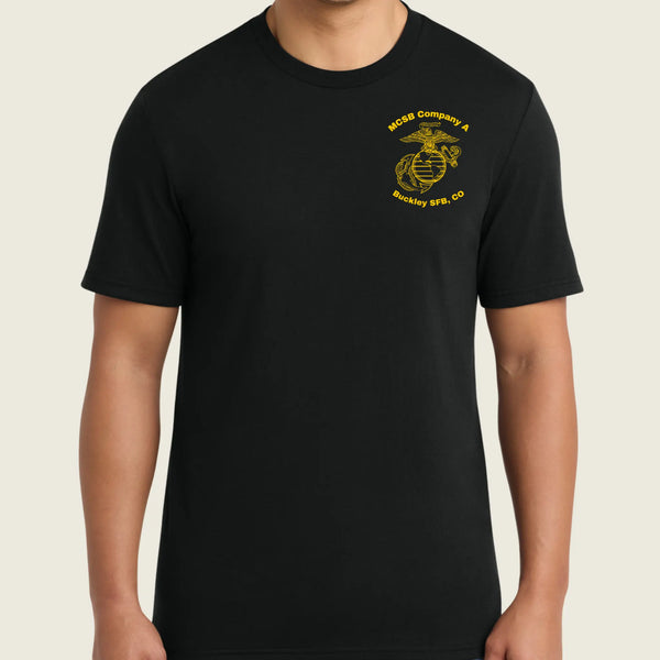 A man wearing a black short-sleeve crewneck t-shirt with a yellow embroidered USMC (United States Marine Corps) Eagle, Globe, and Anchor insignia on the left chest, set against a solid light beige background.