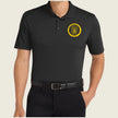 A man wearing a black short-sleeve polo shirt with a yellow circular 'A' emblem on the left chest, paired with black trousers and a black belt, against a light beige background.