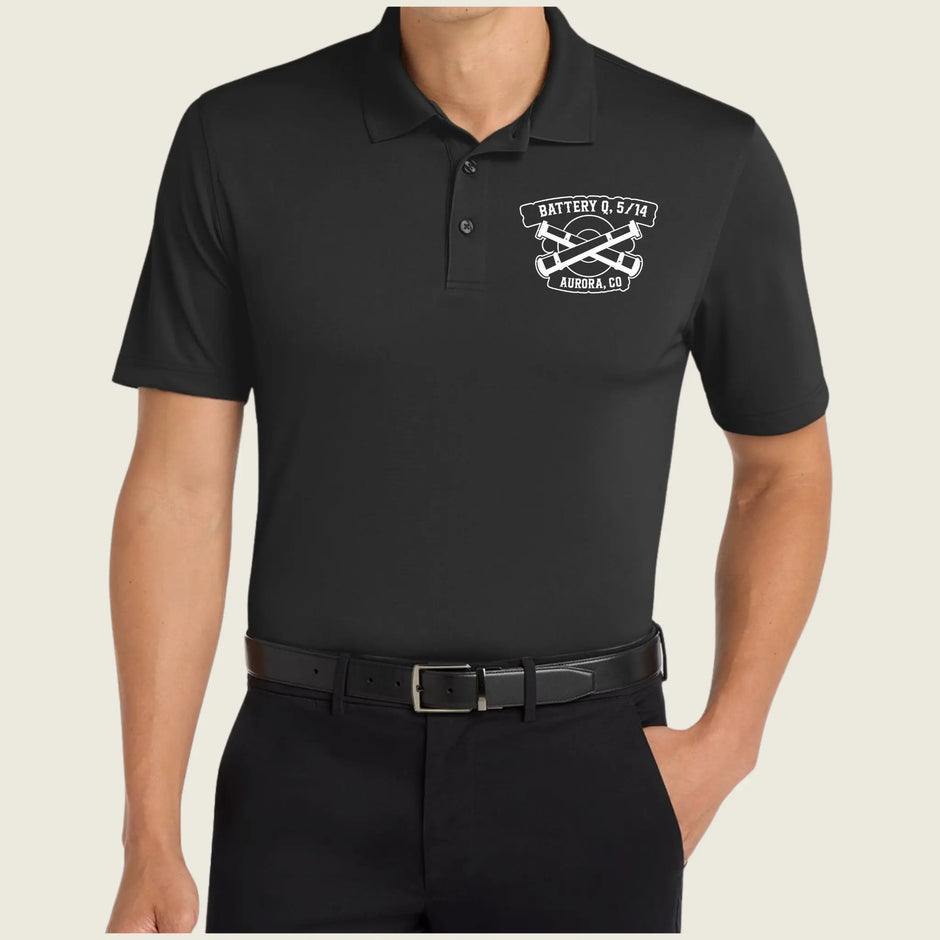 A man wearing a black short-sleeve polo shirt with a white battery and crossed tools graphic on the left chest, paired with black pants and a black belt, against a light beige background.