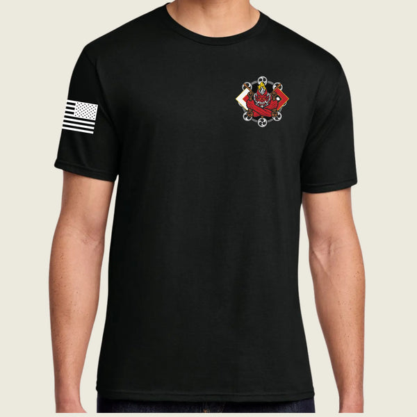 A man wearing a black short-sleeve crewneck t-shirt. The shirt features a small red, white, and black crest graphic on the left chest and a distressed black-and-white American flag graphic on the right sleeve, set against a solid light beige background.