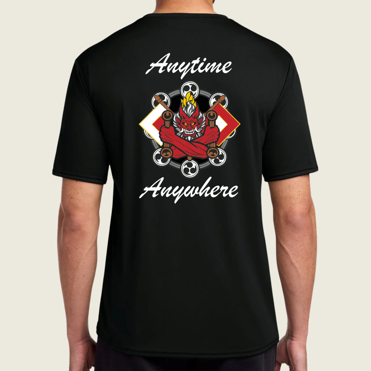 A man wearing a black short-sleeve t-shirt, viewed from the back. The shirt features a large graphic print in the center with a red demon face, soccer balls, and the words 'Anytime Anywhere' in white script.