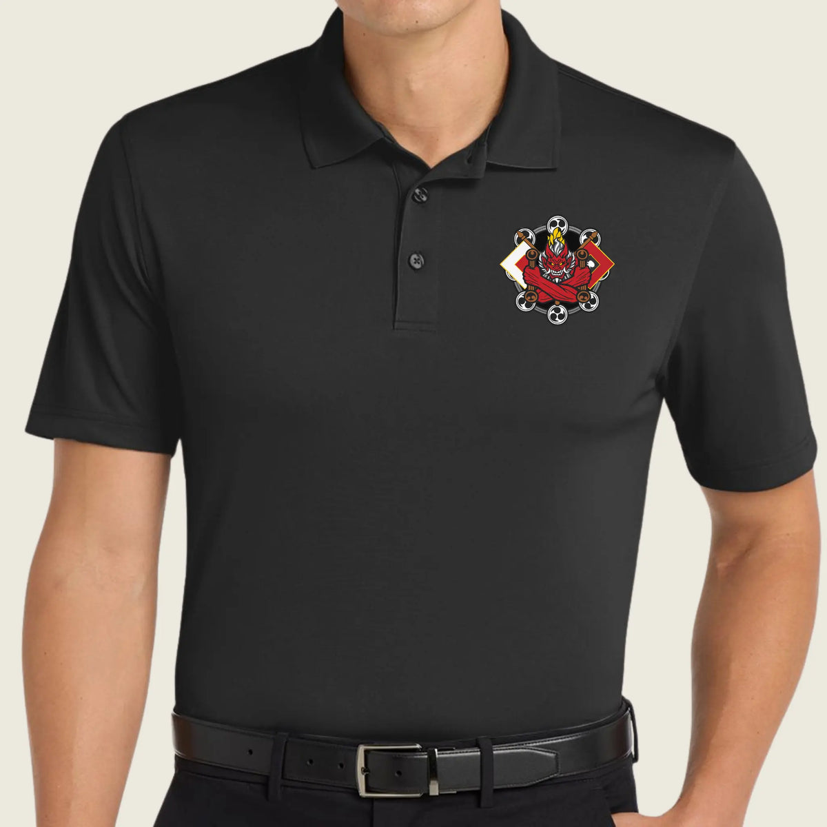 A man wearing a black short-sleeve polo shirt with a small red, white, and black embroidered crest on the left chest, paired with a black belt against a light beige background.