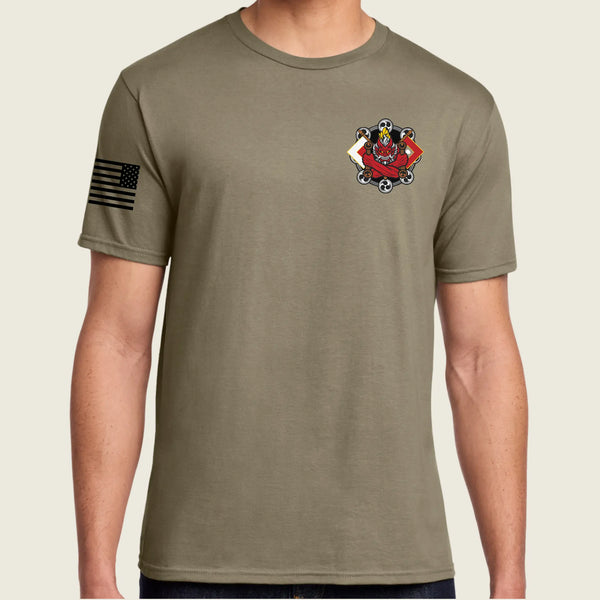 A man wearing an tan short-sleeve t-shirt with a black American flag graphic on the right sleeve and a red and black military-style crest on the left chest, shown against a light beige background.
