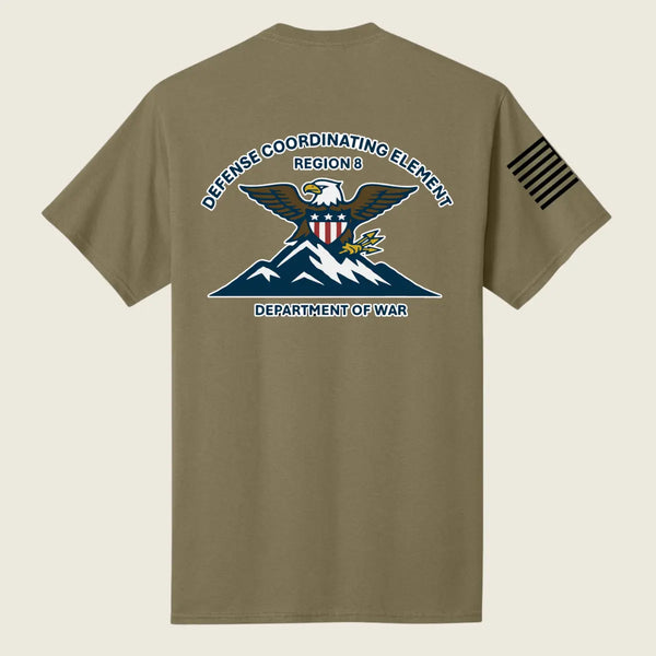 An coyote brown short-sleeve t-shirt shown from the back, featuring a large military-style graphic of an eagle over mountains with the text 'DEFENSE COORDINATING ELEMENT REGION 8' and 'DEPARTMENT OF WAR'. A black striped flag-style patch is printed on the left sleeve.