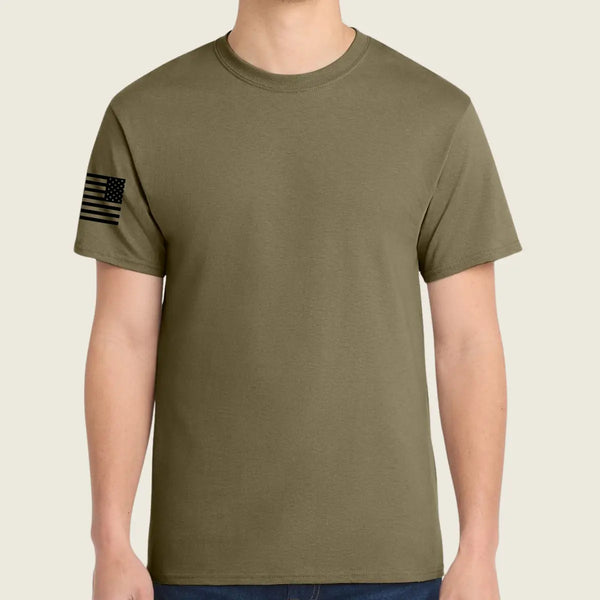 An coyote brown t-shirt worn by a model, featuring a black distressed American flag graphic on the left sleeve, set against a solid light beige background.