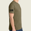 A man wearing a coyote brown t-shirt with a black distressed American flag graphic on the left sleeve, shown from a side profile view against a light beige back