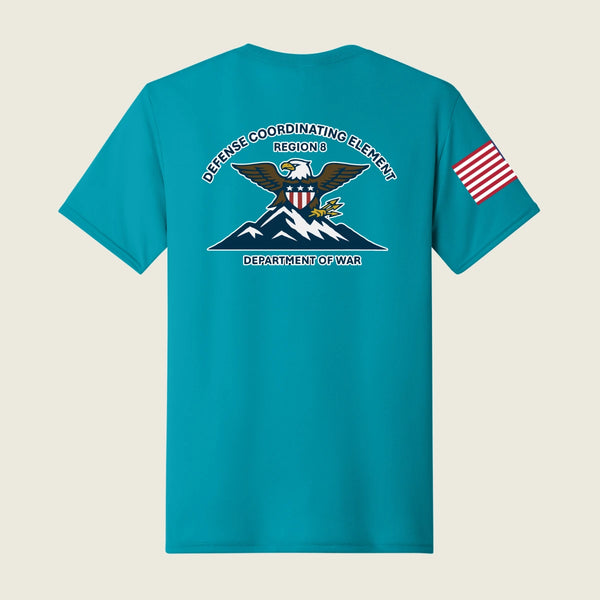 A teal short-sleeve t-shirt shown from the back, featuring a large US Department of War Defense Coordinating Element graphic on the center back and a small American flag print on the left sleeve, set against a light beige background.
