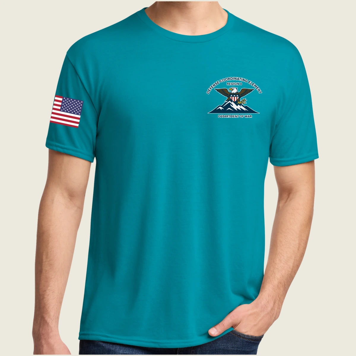 A man wearing a teal short-sleeve t-shirt with a crest on the left chest and an American flag on the right sleeve, shown against a light beige background.