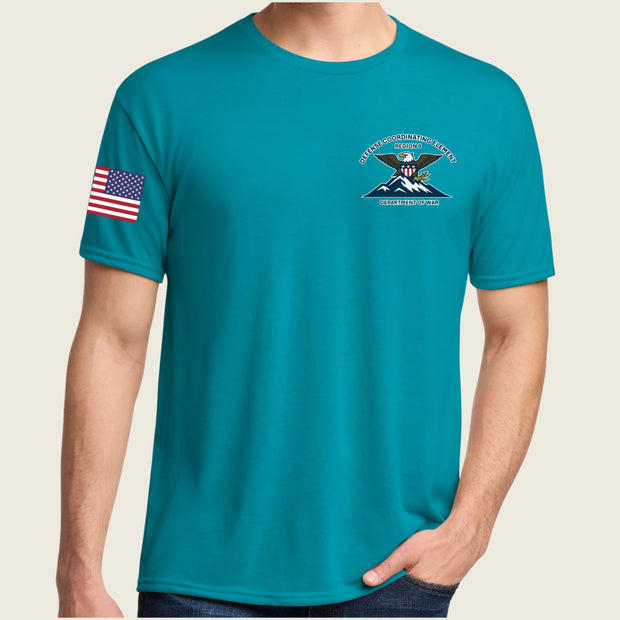 A man wearing a teal short-sleeve t-shirt with a crest on the left chest and an American flag on the right sleeve, shown against a light beige background.
