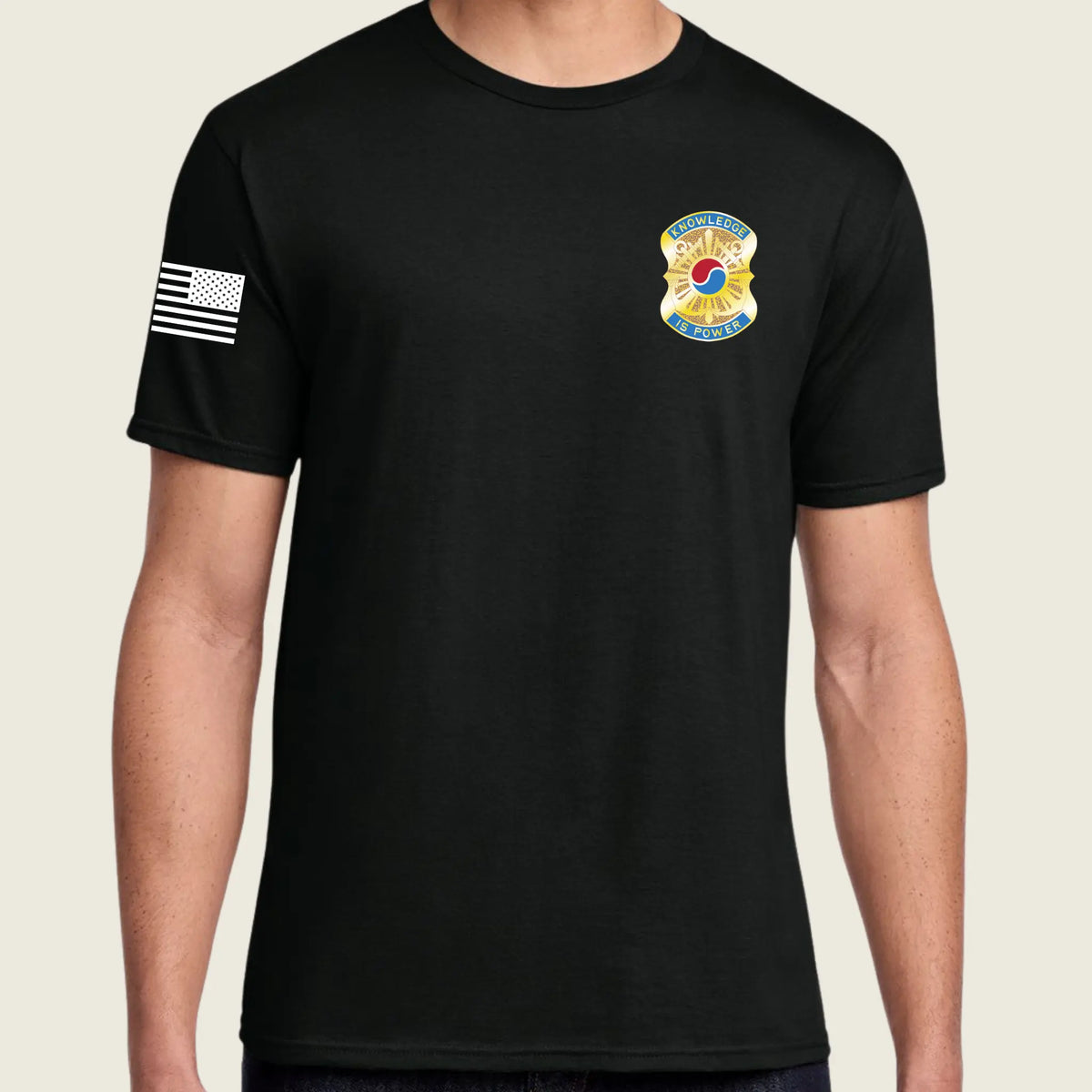 A man wearing a black short-sleeve t-shirt with a white American flag patch on the right sleeve and a yellow and blue fire department badge on the left chest, shown against a light beige background.