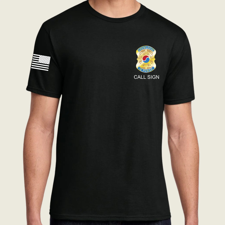 A man wearing a black short-sleeve t-shirt with a white American flag printed on the right sleeve and a yellow and blue fire department style badge graphic with the text 'CALL SIGN' on the left chest, set against a light beige background.