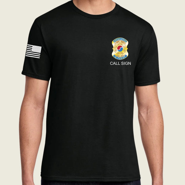 A man wearing a black short-sleeve t-shirt with a white American flag printed on the right sleeve and a yellow and blue fire department style badge graphic with the text 'CALL SIGN' on the left chest, set against a light beige background.
