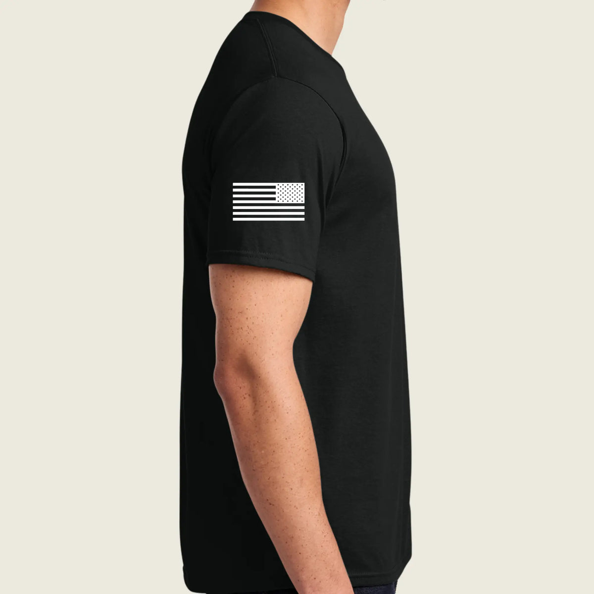 A man wearing a black short-sleeve t-shirt with a white distressed American flag graphic printed on the left sleeve, shown in a side profile view against a light beige background.