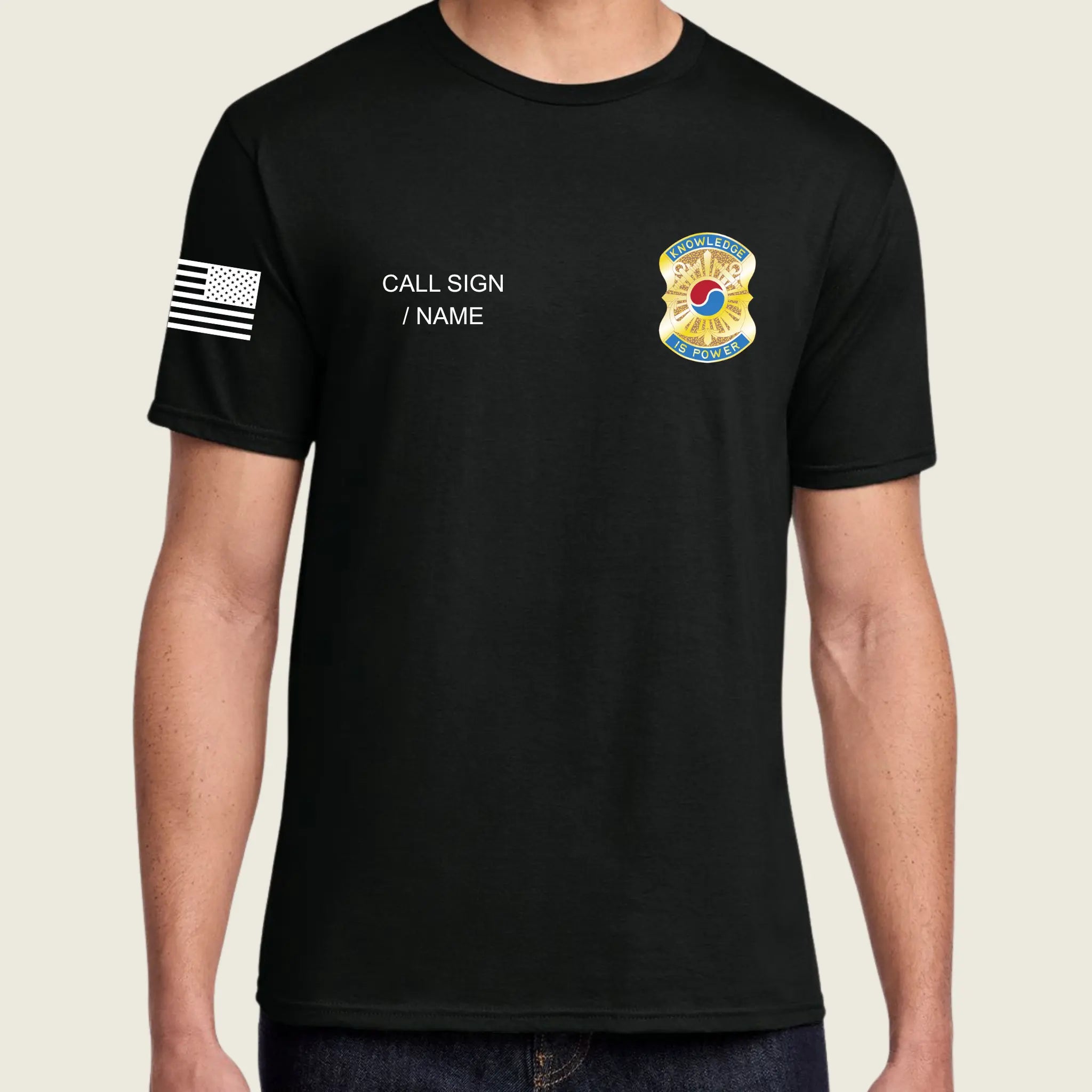 A man wearing a black short-sleeve t-shirt with a black-and-white American flag graphic on the right sleeve, white placeholder text reading 'CALL SIGN / NAME' on the right chest, and a colorful badge graphic on the left chest, shown against a light beige background.