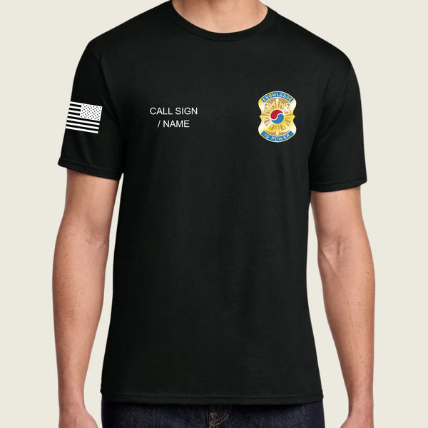A man wearing a black short-sleeve t-shirt with a black-and-white American flag graphic on the right sleeve, white placeholder text reading 'CALL SIGN / NAME' on the right chest, and a colorful badge graphic on the left chest, shown against a light beige background.