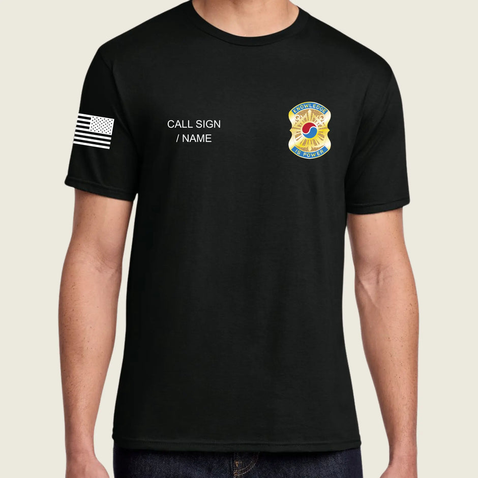 A man wearing a black short-sleeve t-shirt with a black-and-white American flag graphic on the right sleeve, white placeholder text reading 'CALL SIGN / NAME' on the right chest, and a colorful badge graphic on the left chest, shown against a light beige background.