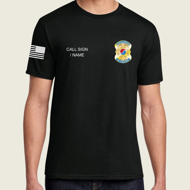 A man wearing a black short-sleeve t-shirt with a black-and-white American flag graphic on the right sleeve, white placeholder text reading 'CALL SIGN / NAME' on the right chest, and a colorful badge graphic on the left chest, shown against a light beige background.