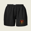 Black athletic shorts with an elastic waistband and a military-style crest patch on the lower left leg, displayed against a light beige background.