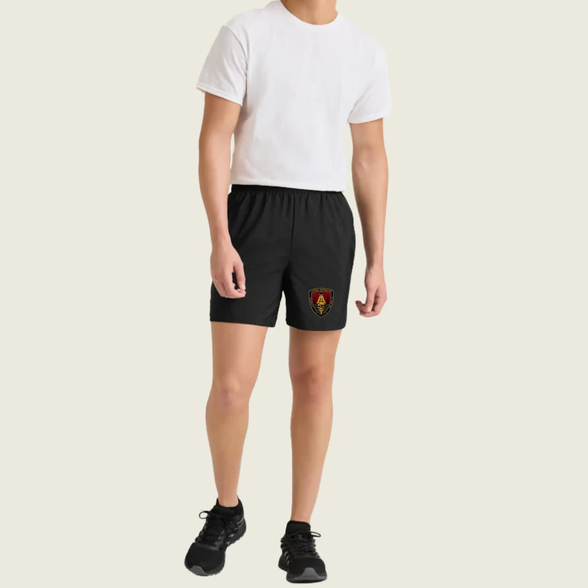 A model wearing black athletic shorts with a small red and yellow crest logo on the left leg, paired with a white t-shirt and black sneakers, against a solid light beige background.