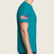 A man wearing a teal short-sleeve t-shirt with a small American flag printed on the left sleeve, shown from a side profile against a light beige background.