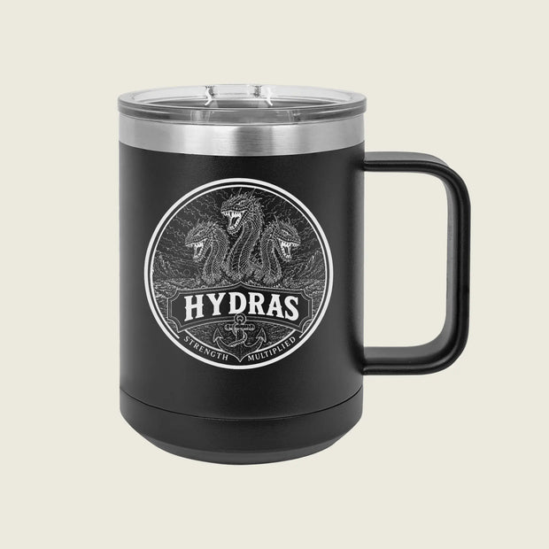 A black insulated travel mug with a clear lid and a black handle, featuring a circular silver graphic of three serpents and the word 'HYDRAS' on the front, set against a light beige background.