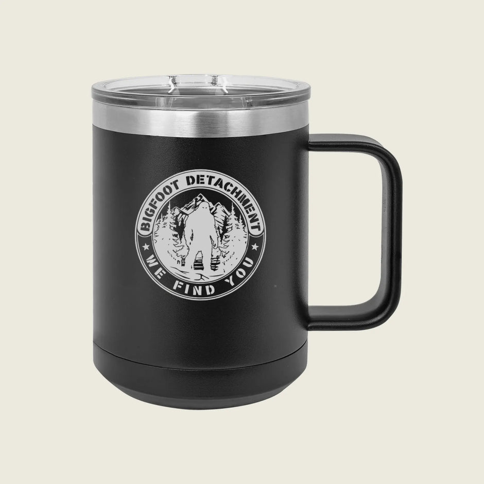 A matte black insulated travel mug with a clear lid and black handle, featuring a circular white graphic that reads 'BIGFOOT DETACHMENT WE FIND YOU' around an illustrated figure in a forest.