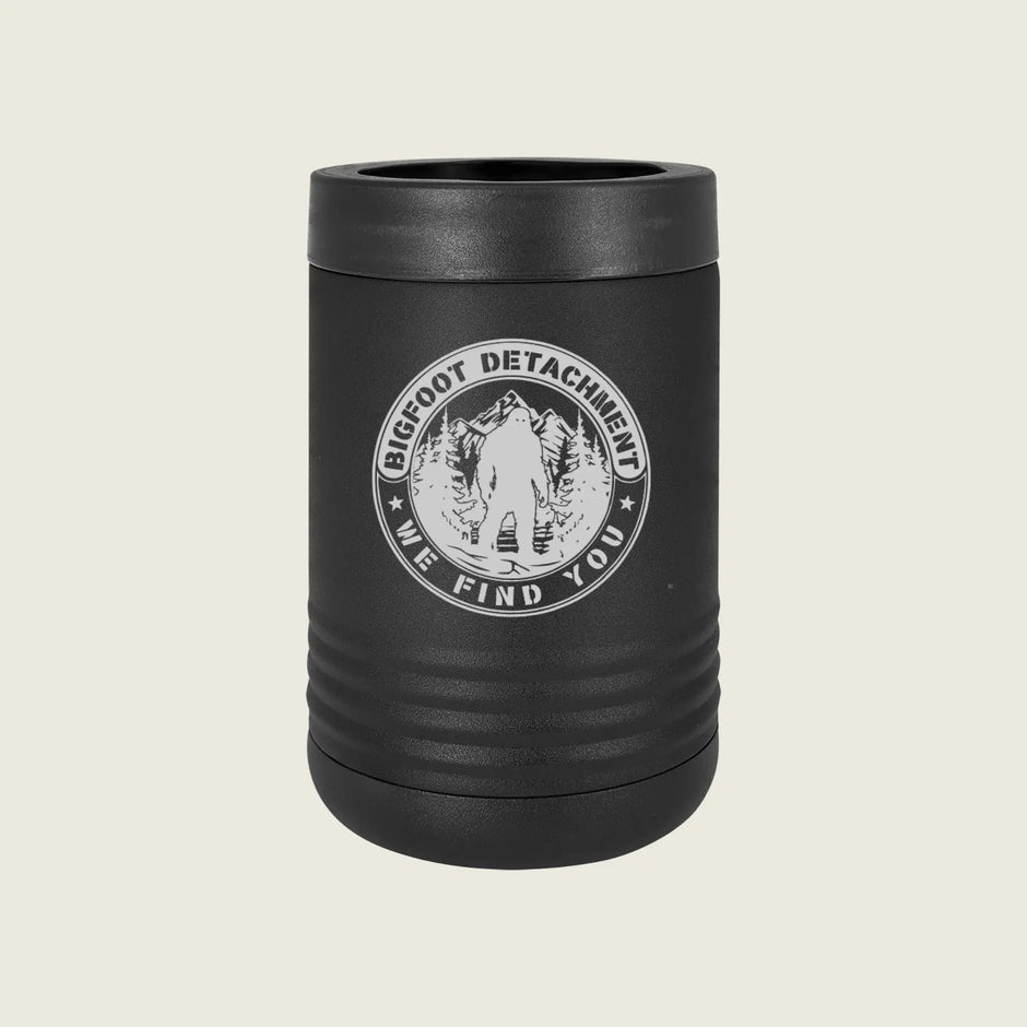 A matte black can cooler with a ribbed design and a white circular 'Bigfoot Detachment We Find You' emblem on the front, set against a solid light beige background.