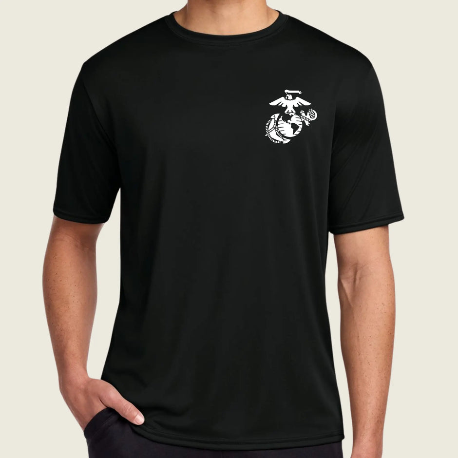A man wearing a black short-sleeve crewneck t-shirt with a small white US Marine Corps Eagle, Globe, and Anchor emblem printed on the left chest, set against a solid light beige background.