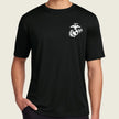 A man wearing a black short-sleeve crewneck t-shirt with a small white US Marine Corps Eagle, Globe, and Anchor emblem printed on the left chest, set against a solid light beige background.