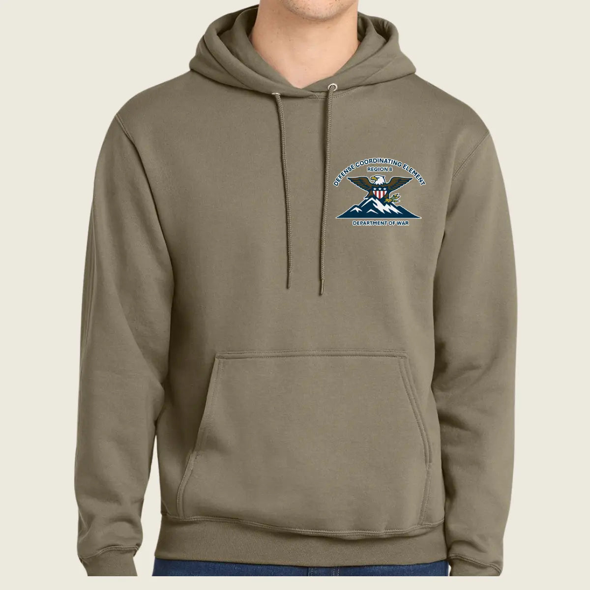 A person wearing a muted olive-brown pullover hoodie with a drawstring hood and kangaroo pocket, featuring a small embroidered United States Army Special Forces crest on the left chest, against a plain light beige background.