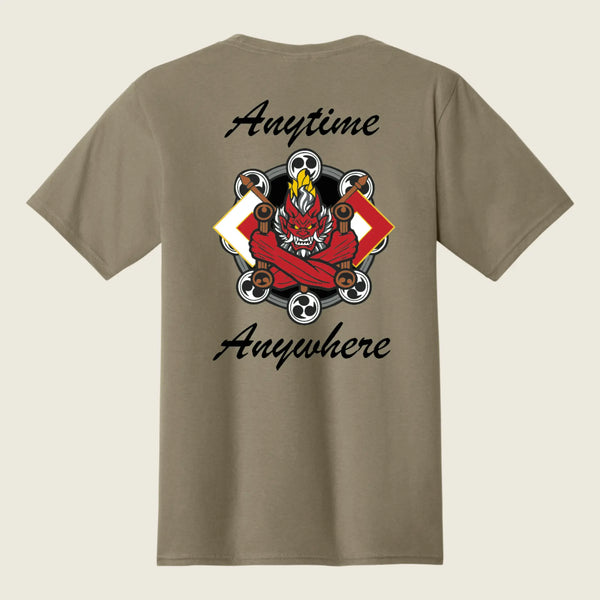 A taupe brown short-sleeve t-shirt shown from the back, featuring a graphic of a red demon skateboarder with flames and skulls, framed by the words 'Anytime Anywhere' in black script.