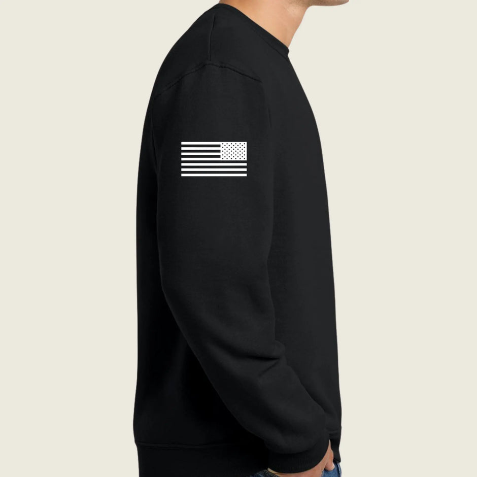 A person wearing a black crewneck sweatshirt, shown from the side. The sweatshirt features a white distressed American flag graphic printed on the upper left sleeve.