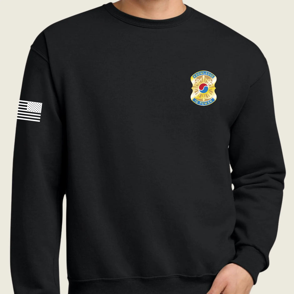 A black crewneck sweatshirt worn by a model, featuring a United States Army emblem on the left chest and a white American flag graphic on the right sleeve, set against a solid light beige background.