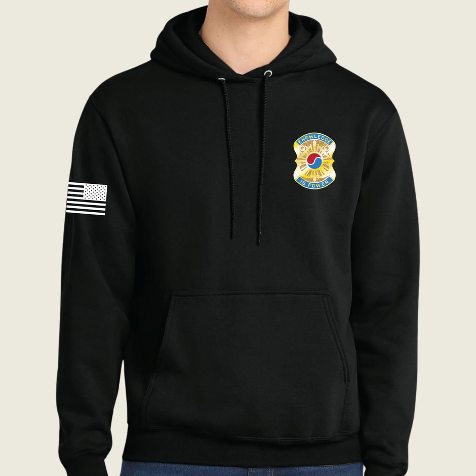 A black pullover hoodie with a drawstring hood and kangaroo pocket, featuring a yellow and blue army emblem on the left chest and a black-and-white American flag patch on the right sleeve, shown on a model against a light beige background.