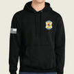 A black pullover hoodie with a drawstring hood and kangaroo pocket, featuring a yellow and blue army emblem on the left chest and a black-and-white American flag patch on the right sleeve, shown on a model against a light beige background.