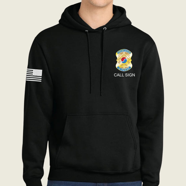 A black pullover hoodie worn by a model, featuring a gold and blue army badge graphic with the text 'CALL SIGN' on the left chest, and a black-and-white American flag patch on the right sleeve.