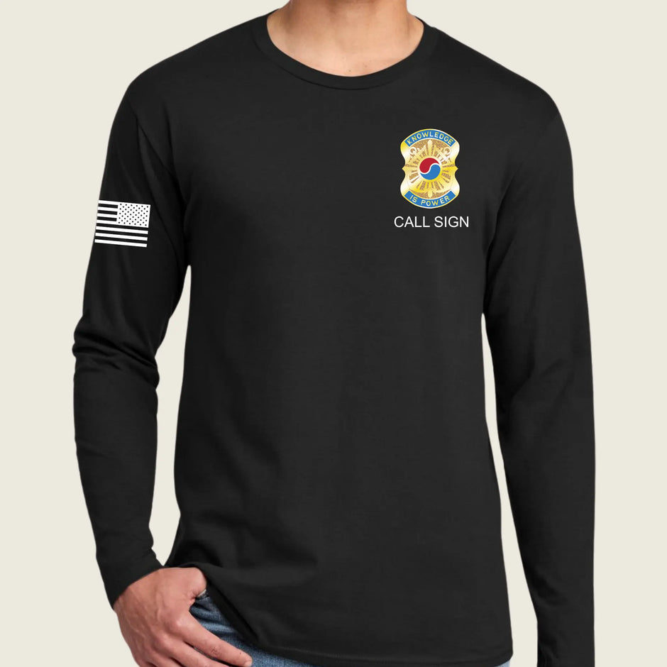 A man wearing a black long-sleeve t-shirt with a US Army emblem on the left chest, 'CALL SIGN' text below it, and a black and white American flag on the right sleeve, set against a light beige background.